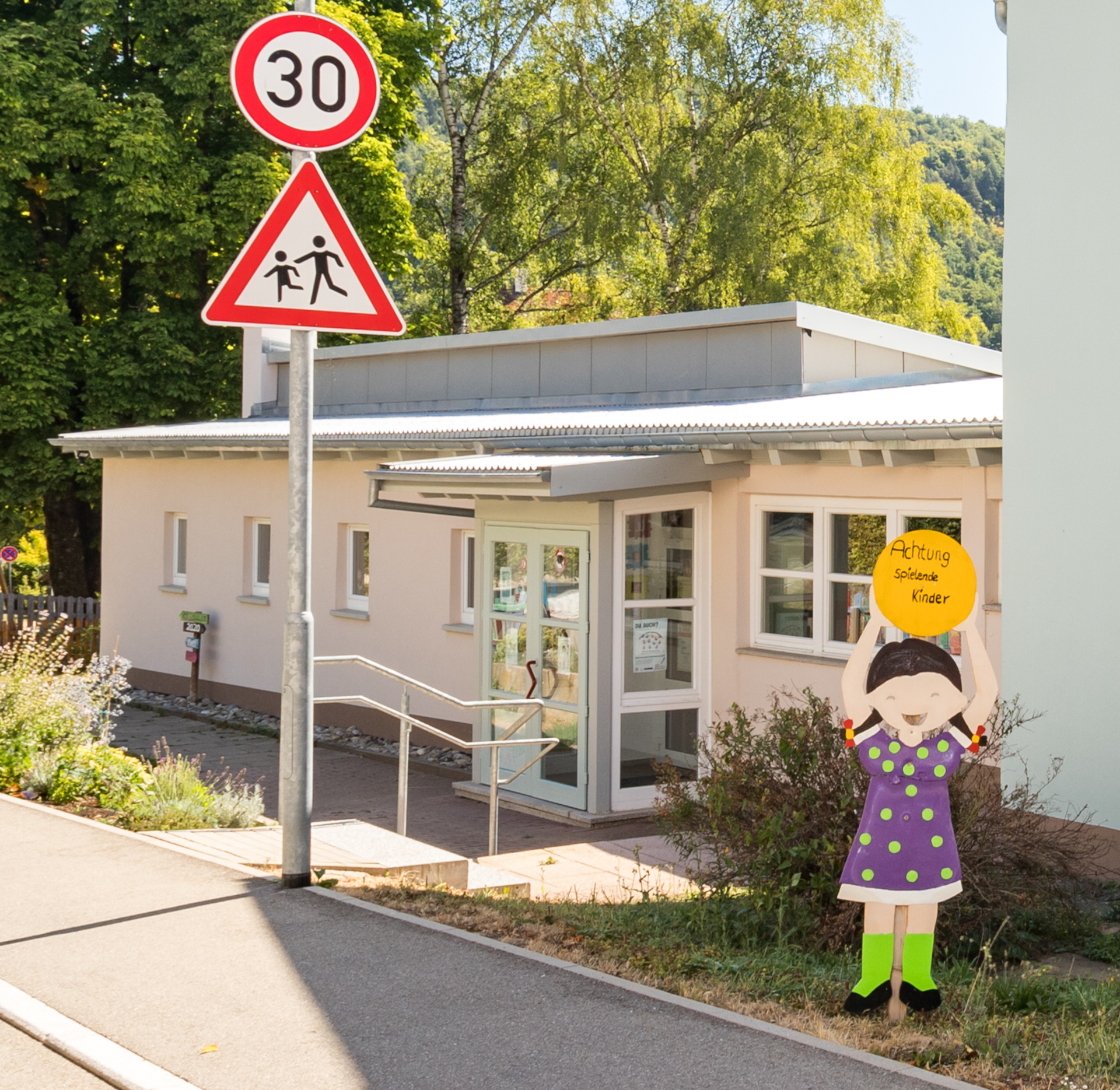 Eingangsbereich mit Hinweisschild auf spielende Kinder und Geschwindigkeitsbegrenzung 30km/h. Rechts eine Mädchenfigur mit Hinweis auf spielende Kinder.
