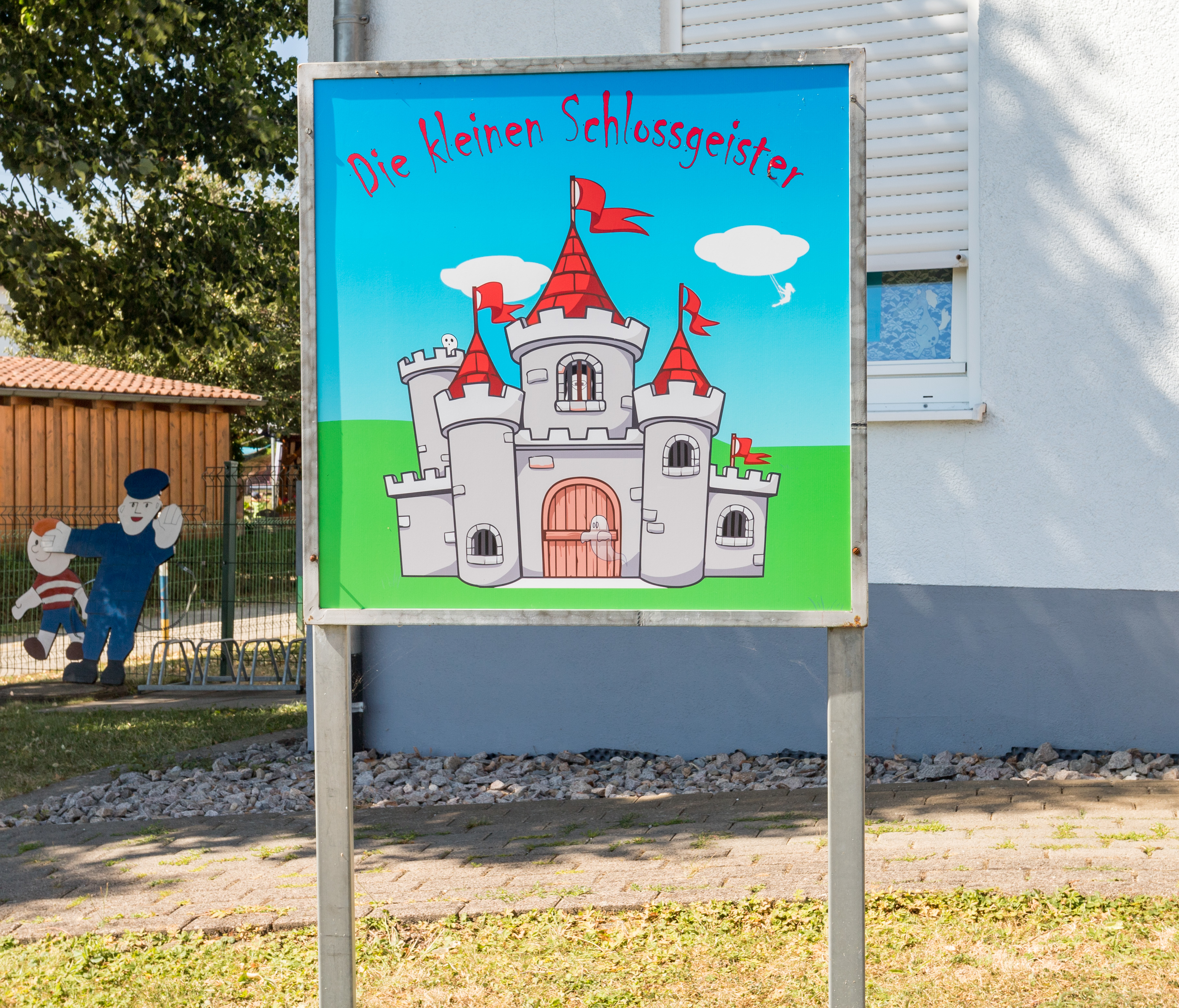 Zum Eingang des Kindergartens weißt ein Schild mit der Aufschrift "Die kleinen Schloßgeister".  Im Hintergrund am Zaun ist aus Holz ein Polizist mit erhobener Hand und ein kleiner Junge angebracht.
