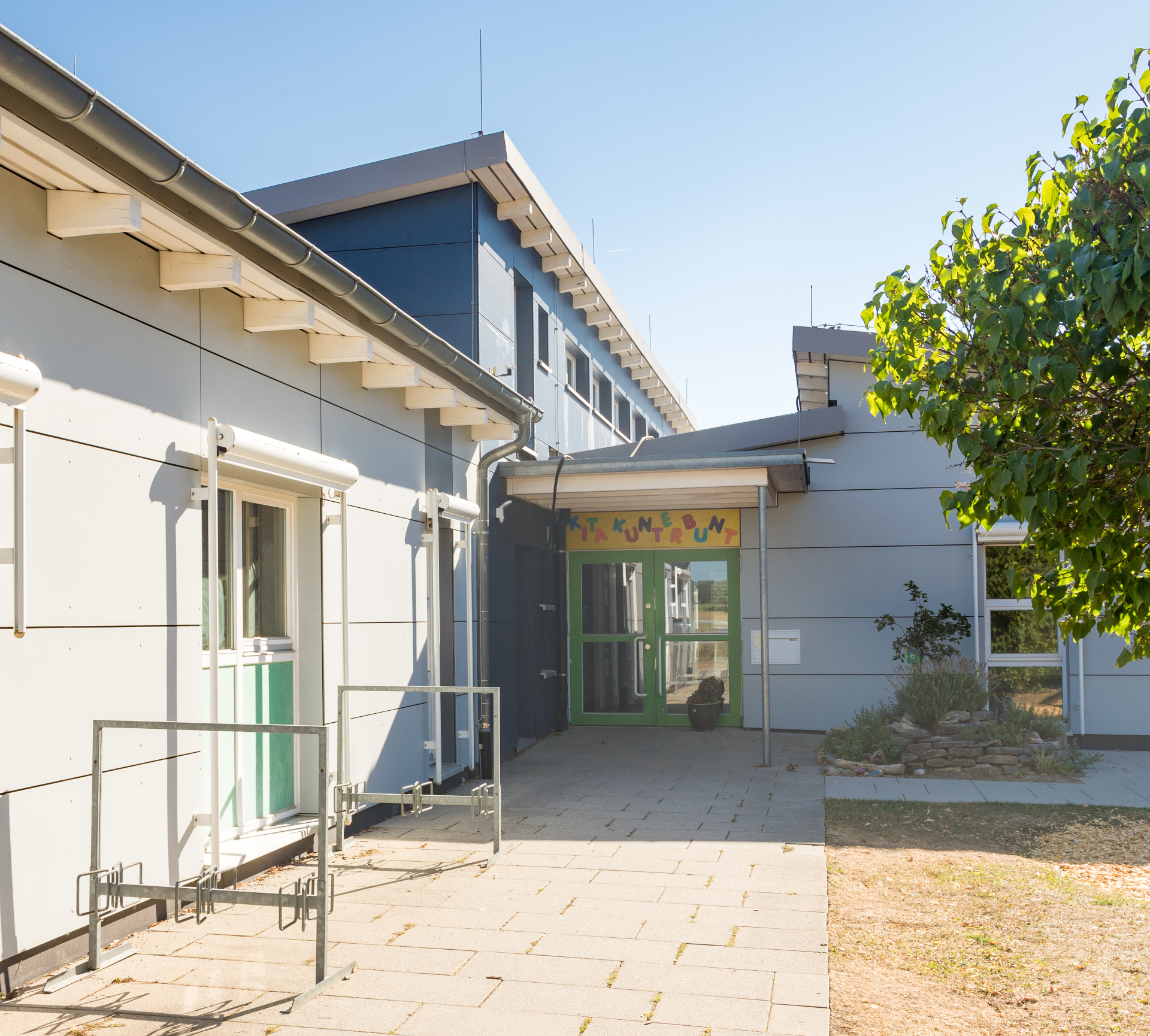 Der Eingangsbereich des Kindergartens ist gepflastert. An der Hauswand steht ein Fahrradständer. Der Eingang ist überdacht. In diesem Bereich steht ein Blumenkübel. Rechts an der Hauswand steht ein Busch. Es ist eine Rasenfläche und ein größerer Baum zu erkennen. 